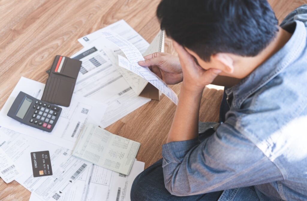 A person sits on the floor, studying a pile of bills and financial statements laid out in front of them. A calculator and notepad sit on top of the pile. The person is rubbing their face with one hand, clearly stressed.