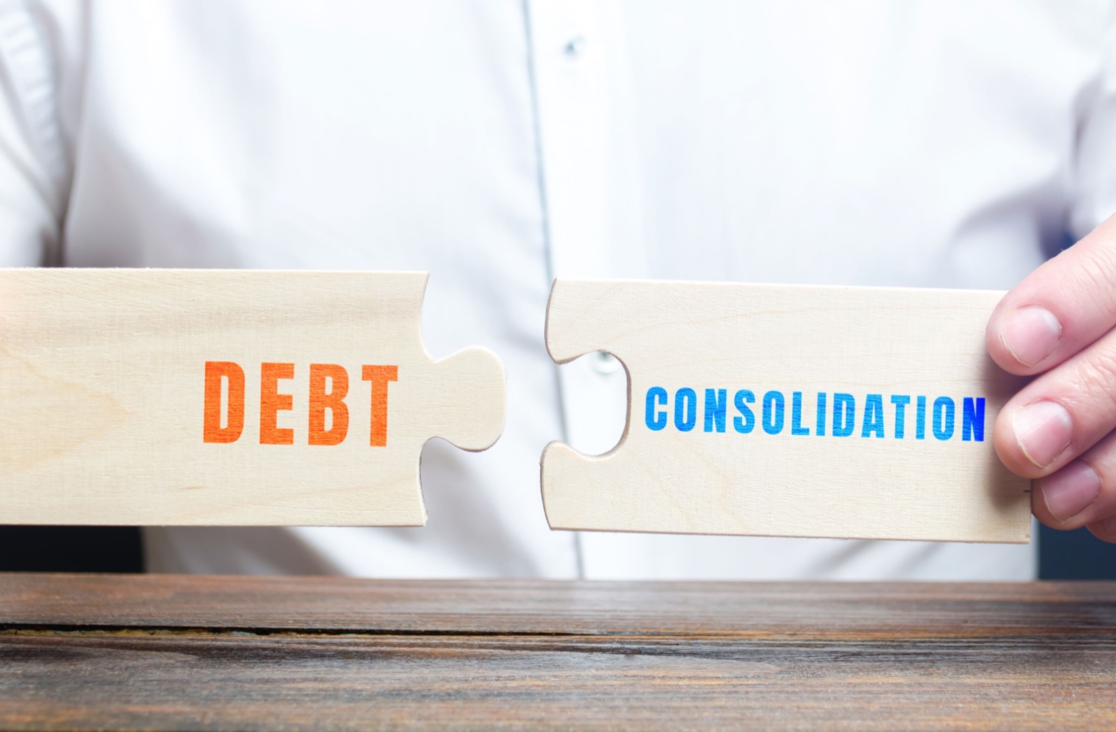 A close-up of a person's hands holding two large wooden puzzle pieces. One says "Debt", and the other says "Consolidation".